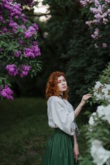Young pretty caucasian woman in romantic outfit walking in blooming spring garden. Artistic outdoor photo of beautiful red-haired girl posing in fragrant lilac blossom.