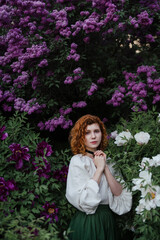Young pretty caucasian woman in romantic outfit walking in blooming spring garden. Artistic outdoor photo of beautiful red-haired girl posing in fragrant lilac blossom.