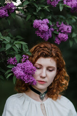 Young pretty caucasian woman in romantic outfit walking in blooming spring garden. Artistic outdoor photo of beautiful red-haired girl posing in fragrant lilac blossom.