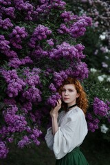 Young pretty caucasian woman in romantic outfit walking in blooming spring garden. Artistic outdoor photo of beautiful red-haired girl posing in fragrant lilac blossom.