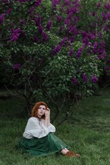 Young pretty caucasian woman in romantic outfit walking in blooming spring garden. Artistic outdoor photo of beautiful red-haired girl posing in fragrant lilac blossom.