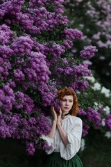 Young pretty caucasian woman in romantic outfit walking in blooming spring garden. Artistic outdoor photo of beautiful red-haired girl posing in fragrant lilac blossom.