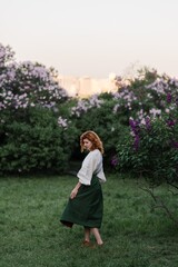 Young pretty caucasian woman in romantic outfit walking in blooming spring garden. Artistic outdoor photo of beautiful red-haired girl posing in fragrant lilac blossom.