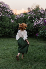 Young pretty caucasian woman in romantic outfit walking in blooming spring garden. Artistic outdoor photo of beautiful red-haired girl posing in fragrant lilac blossom.