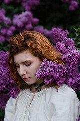 Young pretty caucasian woman in romantic outfit walking in blooming spring garden. Artistic outdoor photo of beautiful red-haired girl posing in fragrant lilac blossom.