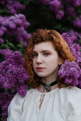 Young pretty caucasian woman in romantic outfit walking in blooming spring garden. Artistic outdoor photo of beautiful red-haired girl posing in fragrant lilac blossom.