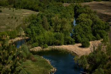 A small winding river flows between grassy banks and fields, blue water and fresh foliage, a...