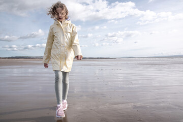 Child in Raincoat Walking on Wet Beach. A young girl in a pale yellow raincoat and pink sneakers...