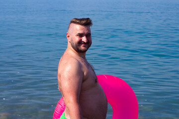 Horizontal photo. Mature fat white man with big belly, stubble, magenta inflatable swim ring,...