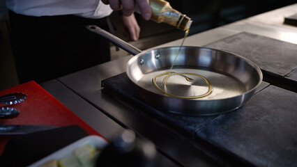 Chef pouring oil into stainless steel pan in restaurant kitchen. Professional chef pouring oil from a bottle into a stainless steel pan on a stove top, preparing to cook in a restaurant kitchen
