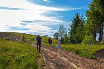 Family Hiking Up Rural Mountain Trail. Three hikers walking up a dirt road through green fields and...