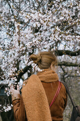 woman with red hair stands in front of a tree with pink flowers. She's wearing a brown coat, scarf.
