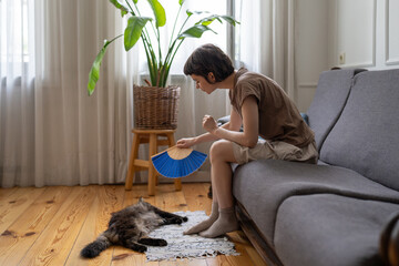 Caring woman fan overheating lazy cat on floor in hot apartment with no AC. Pet owner struggle in...