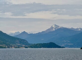 Panoramic view of Lake Como with forested promontory and distant snow-capped Monte Legnone rising over layered alpine ridges under dramatic cloudy sky, Lombardy, northern Italy, tranquil atmosphere