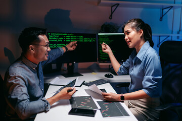 Adult male and female developers analyze stock market data and coding late at night focusing on AI...