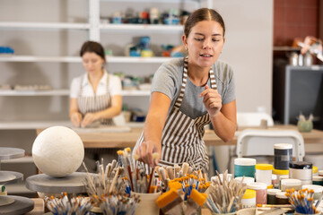 Near place where brushes and paints are stored, student reaches out with her hand to stand with...