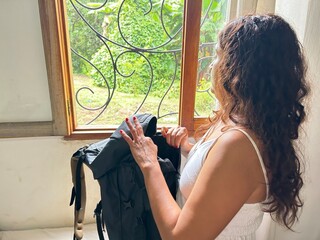 a woman whose face is not visible, with an open backpack, while looking out the window at the green landscape of the tropics, is in the lodge, unpacking her backpack.
