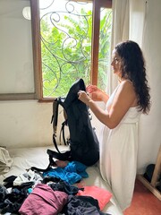 a woman smiles as she unpacks the backpack, on the bed you can see the clothes that were inside, you can see a window, which overlooks the courtyard full of tropical vegetation.