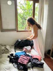 woman unpacking her travel backpack, her hand is inside the backpack, looking for something, her clothes are lying on the bed, the window shows the lush vegetation of the place.