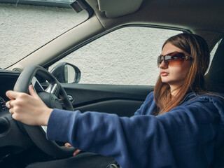 A young inexperienced woman is driving a car with her sunglasses on. She is wearing a blue hoodie....