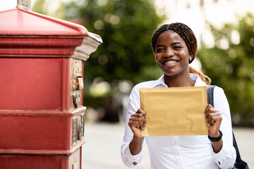 Portrait of smiling black female tourist standing next to mailbox and holding letter.