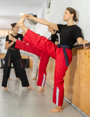 Group of girls in gym are stretching leg muscles, leaning against wall and doing exercises to warm...
