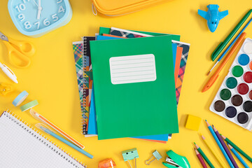 Bright school supplies, stationery, a stack of notebooks in the center of the composition, pencils, a sharpener in the form of an airplane, watercolor paints, an alarm clock placed on a yellow table