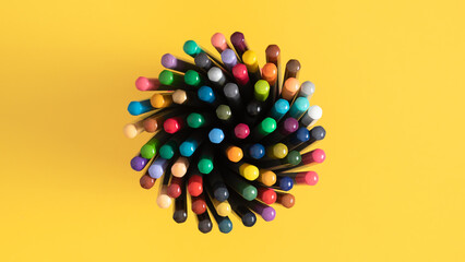 Multicolored pencils on a stand in the center of a yellow table ideal for academic or creative themes. Back to school concept. Banner.