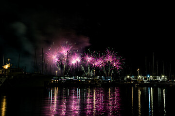 Colorful fireworks exploding over a marina at night, reflected in the calm harbor water with yachts and masts silhouetted under a starless sky with copy space
