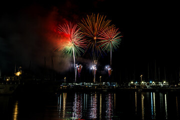 Colorful fireworks exploding over a marina at night, reflected in the calm harbor water with yachts...