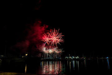 Colorful fireworks exploding over a marina at night, reflected in the calm harbor water with yachts and masts silhouetted under a starless sky with copy space