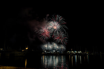 Colorful fireworks exploding over a marina at night, reflected in the calm harbor water with yachts...