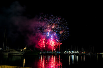 Colorful fireworks exploding over a marina at night, reflected in the calm harbor water with yachts and masts silhouetted under a starless sky with copy space