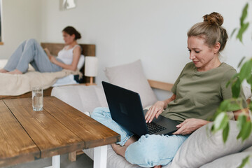 Middle age woman working on laptop at home with another woman relaxing nearby, promoting health and...