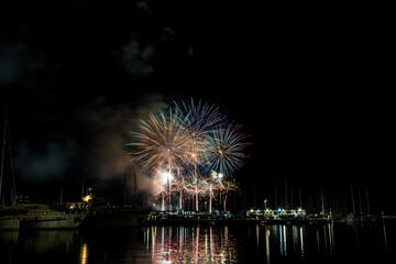 Colorful fireworks exploding over a marina at night, reflected in the calm harbor water with yachts...