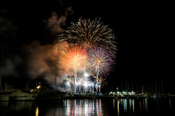 Colorful fireworks exploding over a marina at night, reflected in the calm harbor water with yachts...