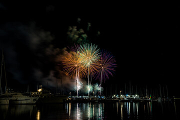 Colorful fireworks exploding over a marina at night, reflected in the calm harbor water with yachts...
