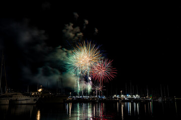 Colorful fireworks exploding over a marina at night, reflected in the calm harbor water with yachts...