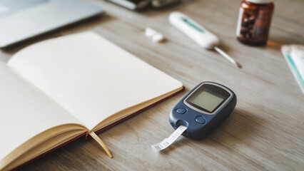 Glucometer with test strip on wooden table, blurred pills and open notebook, concept of diabetes monitoring