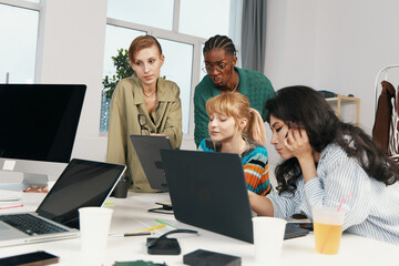 Young adult woman colleagues working together on laptops in modern workplace, socializing and...