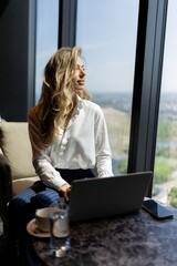 A young woman with blonde hair sits in a stylish and modern workspace, gazing thoughtfully out of a...