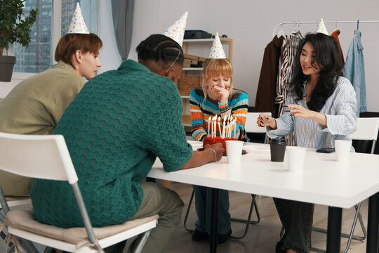 Young adult woman celebrating birthday with business colleagues and coworkers in workplace, wearing party hats and socializing in casual work environment.