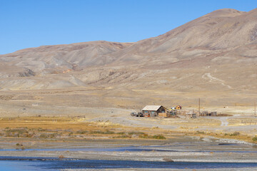 Remote landscape featuring a rustic wooden cabin beside a calm river, surrounded by arid mountains...
