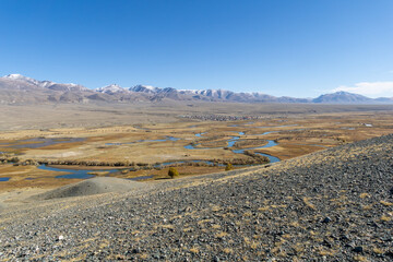 Scenic landscape featuring winding river flowing through golden grasslands, framed by majestic mountains under a clear blue sky, showcasing the beauty of nature and tranquility