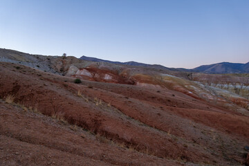 Expansive landscape showcasing vibrant red and earthy tones of rolling hills under a clear sky, highlighting the natural beauty and textures of the terrain at dusk