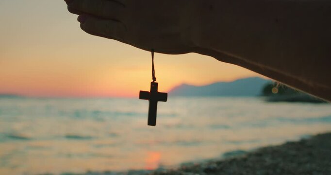 Hand holding a wooden cross necklace in front of the sea during peaceful sunset