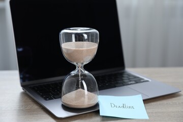 Sticky note with word Deadline, hourglass and laptop on wooden table indoors, closeup