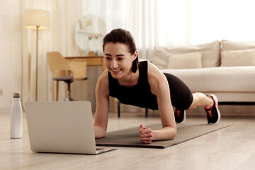 Young woman doing plank exercise during online workout with coach via laptop at home. Sport and...