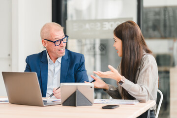 Two businesspeople having lively discussion in modern office with laptop and tablet on table, showing positive and engaged communication