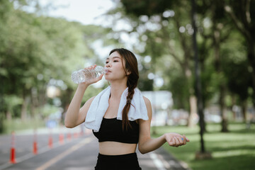 Young Asian woman drinking water after exercise, standing outdoors in workout clothes with towel,...